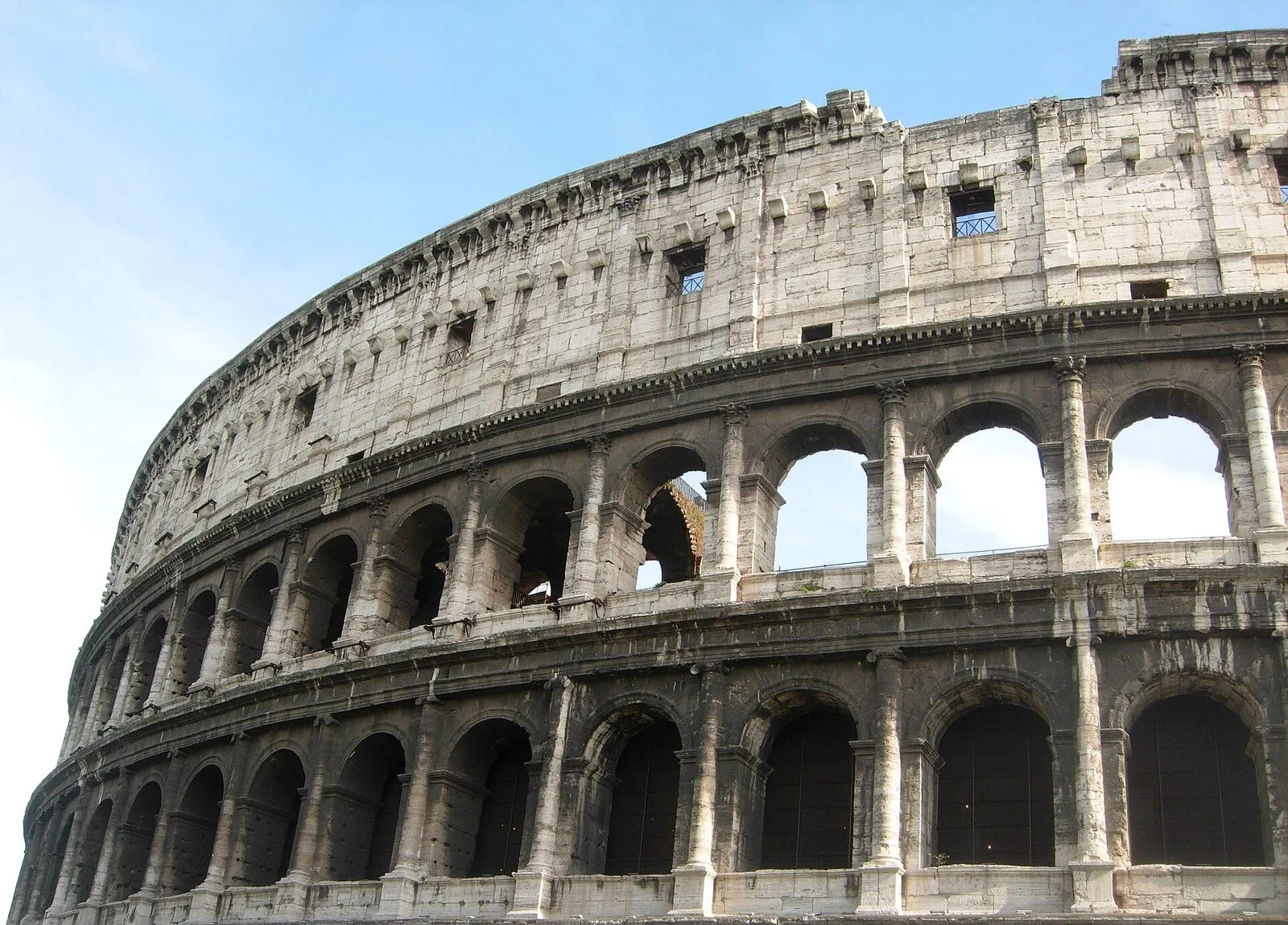 Il fianco esterno del Colosseo visto dal basso contro il cielo azzurro: tre ordini di arcate in travertino con semicolonne e l'attico superiore con le piccole finestre quadrate