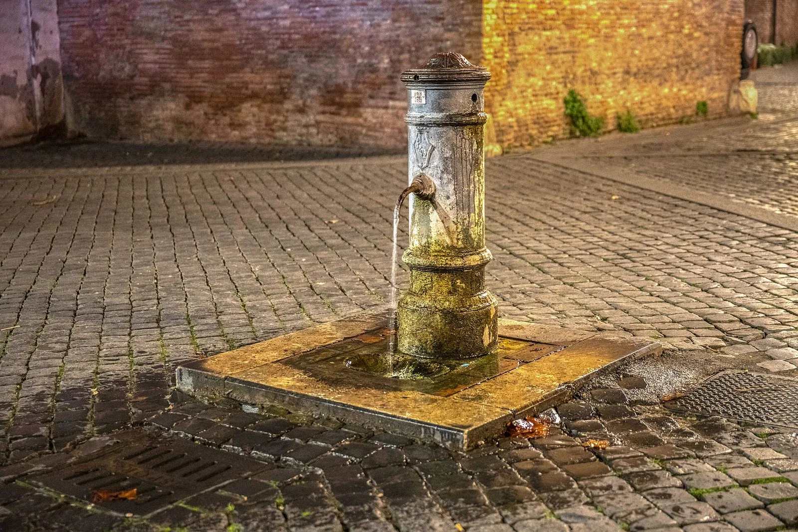 Un nasone, la tipica fontanella romana in ghisa a forma di colonnina, da cui scorre un filo d'acqua sul basamento in pietra, in una piazzetta lastricata di sampietrini con un muro di mattoni sullo sfondo, illuminato dalla luce serale